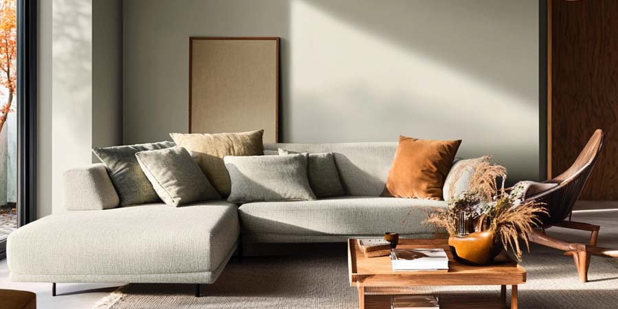 Salon lumineux avec murs en peinture Tilleul Gris Vert, canapé en tissu vert clair, coussins beiges et mobilier bois naturel.